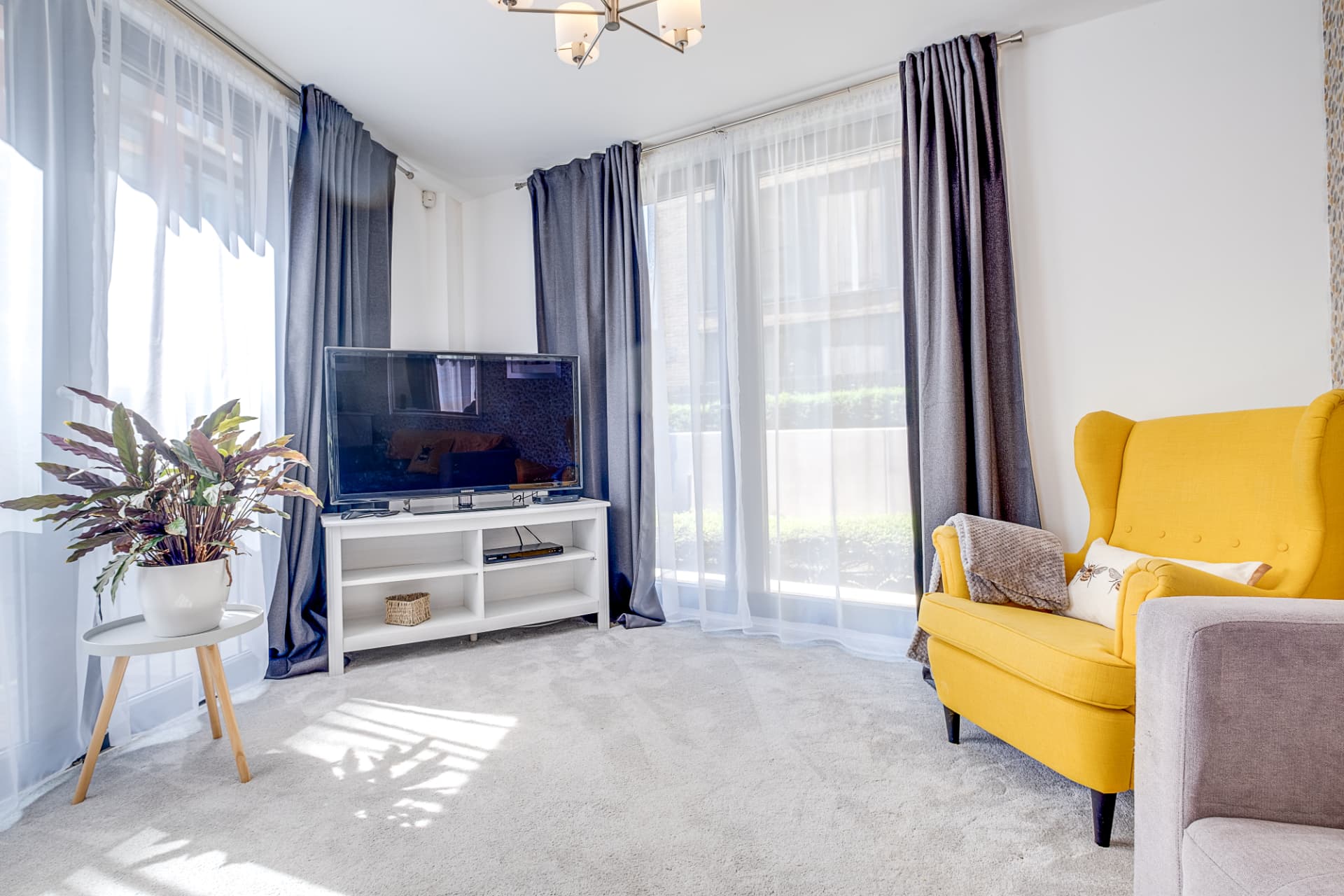 Bright lounge with mustard wing chair, media unit and layered sheer and grey curtains