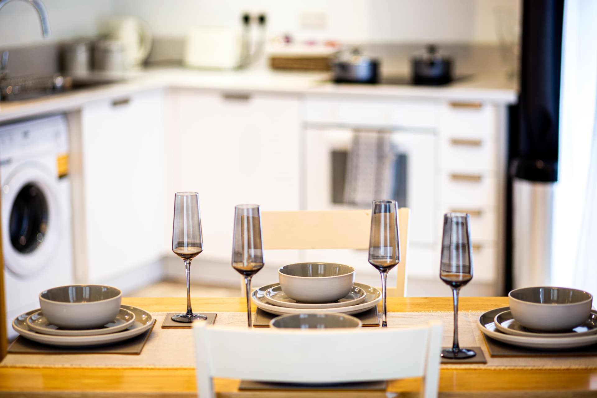 Dining table dressed with glassware and crockery in front of a fitted white kitchen